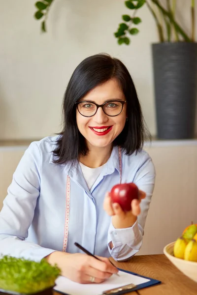 Nutritionist, dietitian woman at the office, hold apple in the hand, healthy vegetables and fruits, healthcare and diet concept. Female nutritionist with fruits working at her desk.