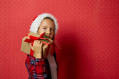 Noel Baba şapkalı pozitif küçük kız elinde Noel hediyesi ve hediye kutusu, kırmızı arka planda izole edilmiş, mutlu yıllar.