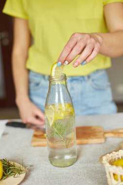 Kadın eli cam şişeye limon koyuyor, kadın hazırlanıyor, limon ve biberiyeyle sağlıklı sudan arınıyor, evde beyaz bir masada taze limonata içiyor, yaz içkisi içiyor.
