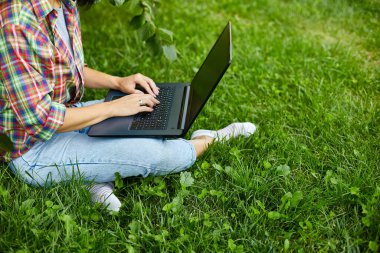 Serbest çalışan, bilgisayarlı bilgisayarı ile parkta çimlerin üzerinde oturan, serbest çalışan, defterle çalışan, dışarıda çalışan, okuyan, online eğitim alan bir kadın. Boşluğu kopyala