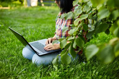 Serbest çalışan, bilgisayarlı bilgisayarı ile parkta çimlerin üzerinde oturan, serbest çalışan, defterle çalışan, dışarıda çalışan, okuyan, online eğitim alan bir kadın. Boşluğu kopyala