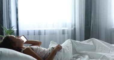 Küçük kız uykusundan uyanır ve telefonla konuşur, büyük ve rahat bir yatak beyaz çarşaf, aile hayatı, rahat yastık, yatak, günaydın
