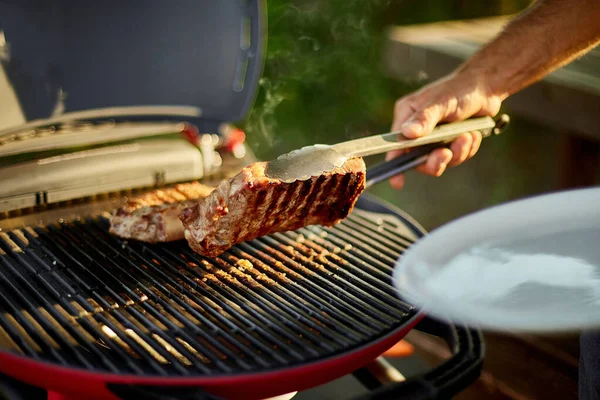 男は木の板にグリルステーキ肉を食べる準備ができて置く 裏庭でバーベキューグリルの屋外でガスグリルで男性ローストステーキ肉 夏の家族のピクニック 自然上の食品 — ストック写真