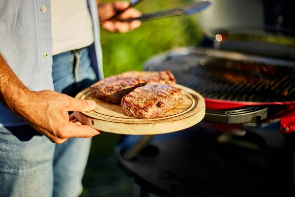 男は木の板にグリルステーキ肉を食べる準備ができて置く 裏庭でバーベキューグリルの屋外でガスグリルで男性ローストステーキ肉 夏の家族のピクニック 自然上の食品 — ストック写真