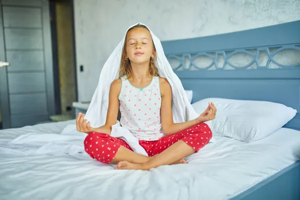 Child girl in pajamas sitting on a bed in lotus pose, Little girl wakes up from sleep on a big and cozy bed white linen in the at home, family lifestyle, comfortable bed, Good morning, Home Family time traditions