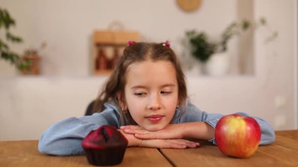 Little girl looking on sweet cake and fresh red apple and choosing in kitchen at home — Stockvideo
