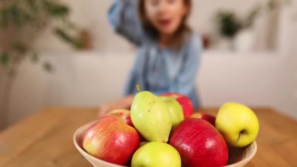 Feliz niña toma una pera del tazón con variedad de frutas en la mesa en casa — Vídeos de Stock