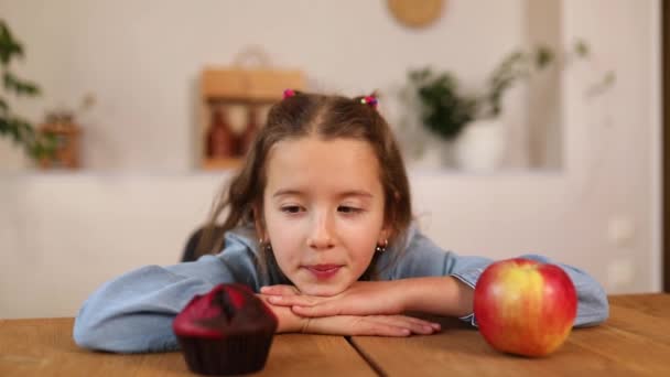 Klein schattig meisje kiezen tussen zoete taart en verse rode appel in de keuken thuis — Stockvideo