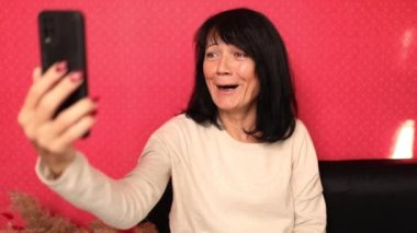 Mutlu gülümseyen olgun bir kadın elinde akıllı telefonuyla arkadaşıyla uzaktan video görüşmesi yapıyor.