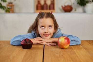 Küçük kız tatlı pastaya ve taze kırmızı elmaya bakıyor ve evde mutfağı seçiyor sağlıklı beslenme alışkanlığı, sağlıksız yemek kavramı.