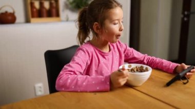 Küçük kız mısır gevreğini sütle yiyor ve akıllı telefondan video izliyor, lezzetli sağlıklı yemekler yiyor, güzel bir çocuğun kahvaltısını yavaş çekimde yapıyor,