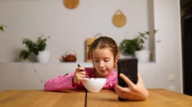 Küçük kız mısır gevreğini sütle yiyor ve akıllı telefondan video izliyor, lezzetli sağlıklı yemekler yiyor, güzel bir çocuğun kahvaltısını yavaş çekimde yapıyor,