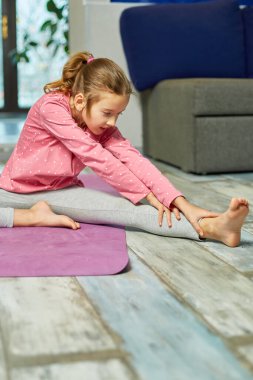 Küçük kız esneme egzersizleri yapıyor, evde fitness minderinde yoga yapıyor spor yapıyor, sağlıklı bir yaşam tarzı sergiliyor. Çocuklar ev karantinası yapıyor