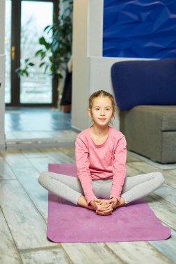 Küçük kız esneme egzersizleri yapıyor, evde fitness minderinde yoga yapıyor spor yapıyor, sağlıklı bir yaşam tarzı sergiliyor. Çocuklar ev karantinası yapıyor