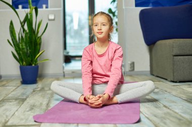 Küçük kız esneme egzersizleri yapıyor, evde fitness minderinde yoga yapıyor spor yapıyor, sağlıklı bir yaşam tarzı sergiliyor. Çocuklar ev karantinası yapıyor