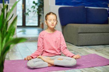 Evde yoga yaparken nilüfer minderinde oturan tatlı küçük kız meditasyon yapıyor spor yapıyor, sağlıklı bir yaşam sürüyor.