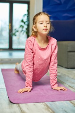 Küçük kız esneme egzersizleri yapıyor, evde fitness minderinde yoga yapıyor spor yapıyor, sağlıklı bir yaşam tarzı sergiliyor. Çocuklar ev karantinası yapıyor