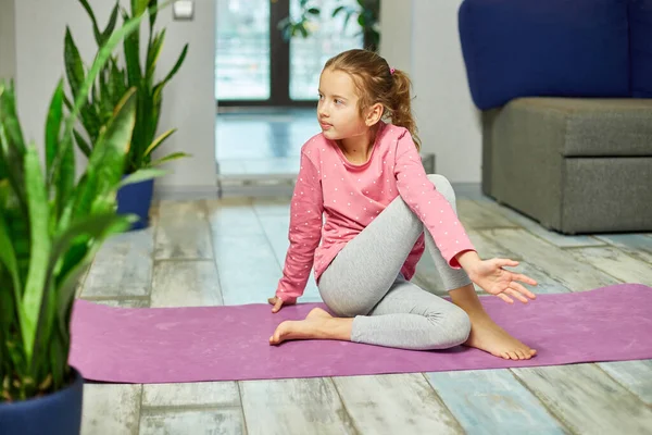 Küçük kız esneme egzersizleri yapıyor, evde fitness minderinde yoga yapıyor spor yapıyor, sağlıklı bir yaşam tarzı sergiliyor. Çocuklar ev karantinası yapıyor