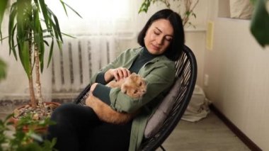 Koltuğunda oturan bir kadın sevimli kızıl kediyi tutuyor, uzun saçlı kediciğine sarılıyor, yeşil evdeki kediyle oynuyor, ev bahçesi kavramı, biyofili tasarımı....