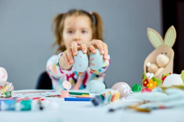Paskalya gününde küçük şirin bir kız çocuğu paskalya yumurtasını elinde tutuyor. Parlak arka planda boyalı yumurtaları olan kız, Mutlu Paskalyalar