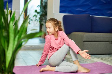 Küçük kız esneme egzersizleri yapıyor, evde fitness minderinde yoga yapıyor spor yapıyor, sağlıklı bir yaşam tarzı sergiliyor. Çocuklar ev karantinası yapıyor