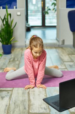 Küçük kız dizüstü bilgisayarda video izliyor ve evde esneme hareketleri yapıyor. Kişisel antrenörle uzak çocuk eğitimi, online eğitim konsepti, spor yapmak, sağlıklı yaşam tarzı.