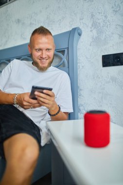 Erkek testi, akıllı telsiz hoparlörü, ses komutlarıyla ev aletlerini kontrol eden adam, evdeki kanepede oturmak, akıllı ev kavramı.