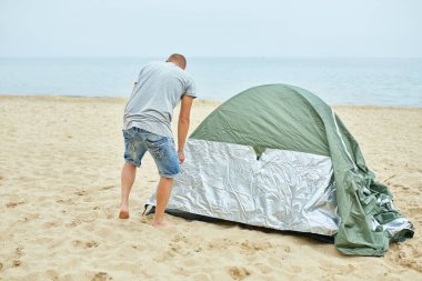 Genç erkek turist sahile yeşil bir çadır kuruyor. Adam yürüyüş için kamp kuruyor, tek başına çadır kuruyor, yeni normal seyahatler yapıyor..