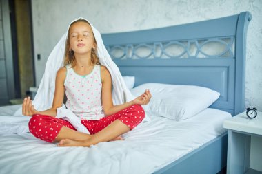 Pijamalı bir kız Lotus pozunda yatağında oturuyor, küçük kız büyük ve rahat bir yatakta uykusundan uyanıyor, beyaz çarşaflar evde, aile hayatı tarzı, rahat bir yatak, günaydın, aile zamanı gelenekleri.