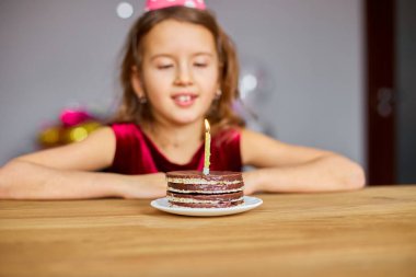 Küçük bir kız doğum günü şapkası takıyor, dilek diliyor, doğum günü pastasına bakıyor, kutlama partisi konsepti için yanan mumlarla..