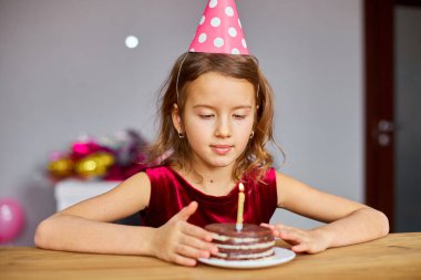 Küçük bir kız doğum günü şapkası takıyor, dilek diliyor, doğum günü pastasına bakıyor, kutlama partisi konsepti için yanan mumlarla..