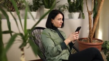 Woman hold mobile phone and surfing internet, type message, sitting on armchair at home garden, Biophilia design.