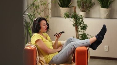 Kulaklıklı, akıllı telefonlu, günlük kıyafetleri olan neşeli bir kadın evdeki koltukta podcast yapıyor. Kadın podcaster canlı yayında, kulüp binasında konuşan kadın..