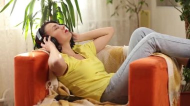 Kulaklıklı kadın, gözleri kapalı müzik dinliyor, podcast turuncu bir kanepede oturuyor. Parlak kıyafetli güzel kadın evde müziğin tadını çıkarıyor.