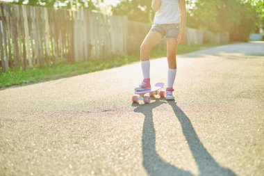 Kaykay kullanmayı öğrenen küçük bir kız, beyaz çoraplar ve pembe ayakkabılar giyen isimsiz bir çocuk yaz şehrinin kırsal kesiminde kaykaya biniyor..