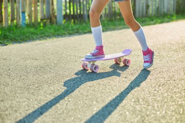 Kaykay kullanmayı öğrenen küçük bir kız, beyaz çoraplar ve pembe ayakkabılar giyen isimsiz bir çocuk yaz şehrinin kırsal kesiminde kaykaya biniyor..