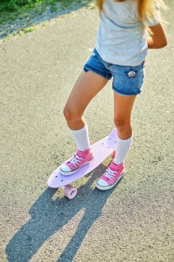 Kaykay kullanmayı öğrenen küçük bir kız, beyaz çoraplar ve pembe ayakkabılar giyen isimsiz bir çocuk yaz şehrinin kırsal kesiminde kaykaya biniyor..