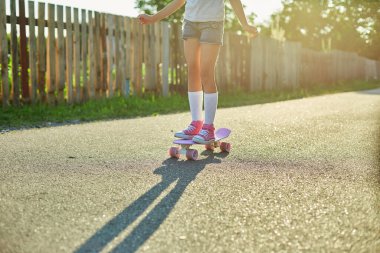 Kaykay kullanmayı öğrenen küçük bir kız, beyaz çoraplar ve pembe ayakkabılar giyen isimsiz bir çocuk yaz şehrinin kırsal kesiminde kaykaya biniyor..