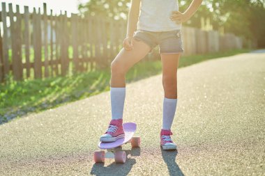 Kaykay kullanmayı öğrenen küçük bir kız, beyaz çoraplar ve pembe ayakkabılar giyen isimsiz bir çocuk yaz şehrinin kırsal kesiminde kaykaya biniyor..