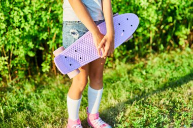 Küçük kız kaykayını tutuyor, beyaz çoraplar ve pembe ayakkabılar giyen anonim bir çocuk yaz şehrinin kırsal kesiminde kaykay sürüyor..