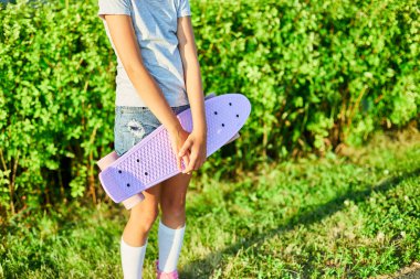 Küçük kız kaykayını tutuyor, beyaz çoraplar ve pembe ayakkabılar giyen anonim bir çocuk yaz şehrinin kırsal kesiminde kaykay sürüyor..