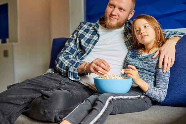 Father and daughter eat popcorn and watch TV film, Dad and child girl watching a movie on a couch at home, happy smiling family spending free time together, relax.