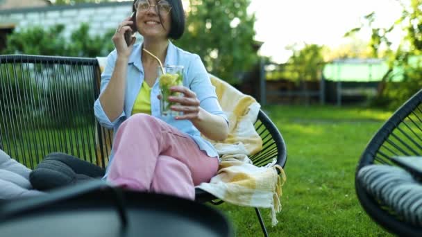 携帯電話で話してガラスの幸せな女性は 自宅で裏庭の屋外で椅子に座って 再利用可能な金属のストローでレモネードを入れ テーブルの上に冷たい飲み物 — ストック動画