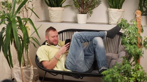 男人用智能手机聊天 躺在家里的沙发上 笑容满面地输入讯息 家庭花园的概念 恋童癖的设计 — 图库视频影像