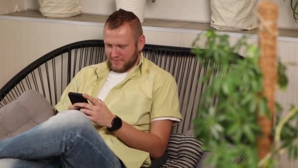 男人用智能手机聊天 坐在家里的扶手椅上 带着快乐的笑容输入讯息 家庭花园 的概念 恋童癖 的设计 — 图库视频影像