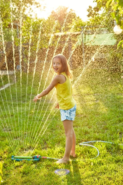 Happy kid girl playing with garden sprinkler run and jump, summer outdoor water fun in the backyard at home, splash on sunny day.