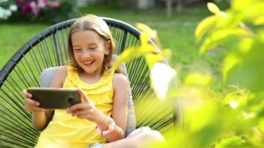 Parkta cep telefonuyla oyun oynayan mutlu bir kız çocuğu ev bahçesinde, arka bahçede akıllı telefon kullanan bir çocuk, güneş ışığı, akıllı telefon bağımlılığı.