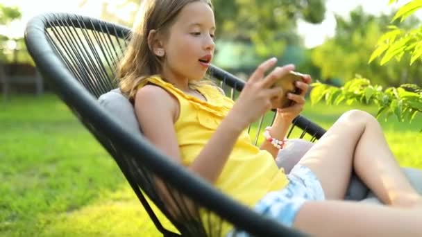 公園の屋外で携帯電話でゲームをプレイする幸せな子供の女の子 自宅の庭 スマートフォン中毒でスマートフォンを使用して子供 — ストック動画