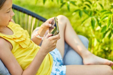 Parkta cep telefonuyla oyun oynayan mutlu bir kız çocuğu ev bahçesinde, arka bahçede akıllı telefon kullanan bir çocuk, güneş ışığı, akıllı telefon bağımlılığı.