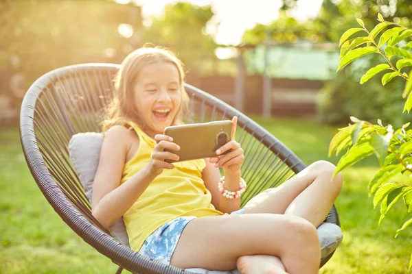 Happy kid girl playing game on mobile phone in the park outdoor, child using smartphone at home garden, backyard, sunlight, smartphone addiction